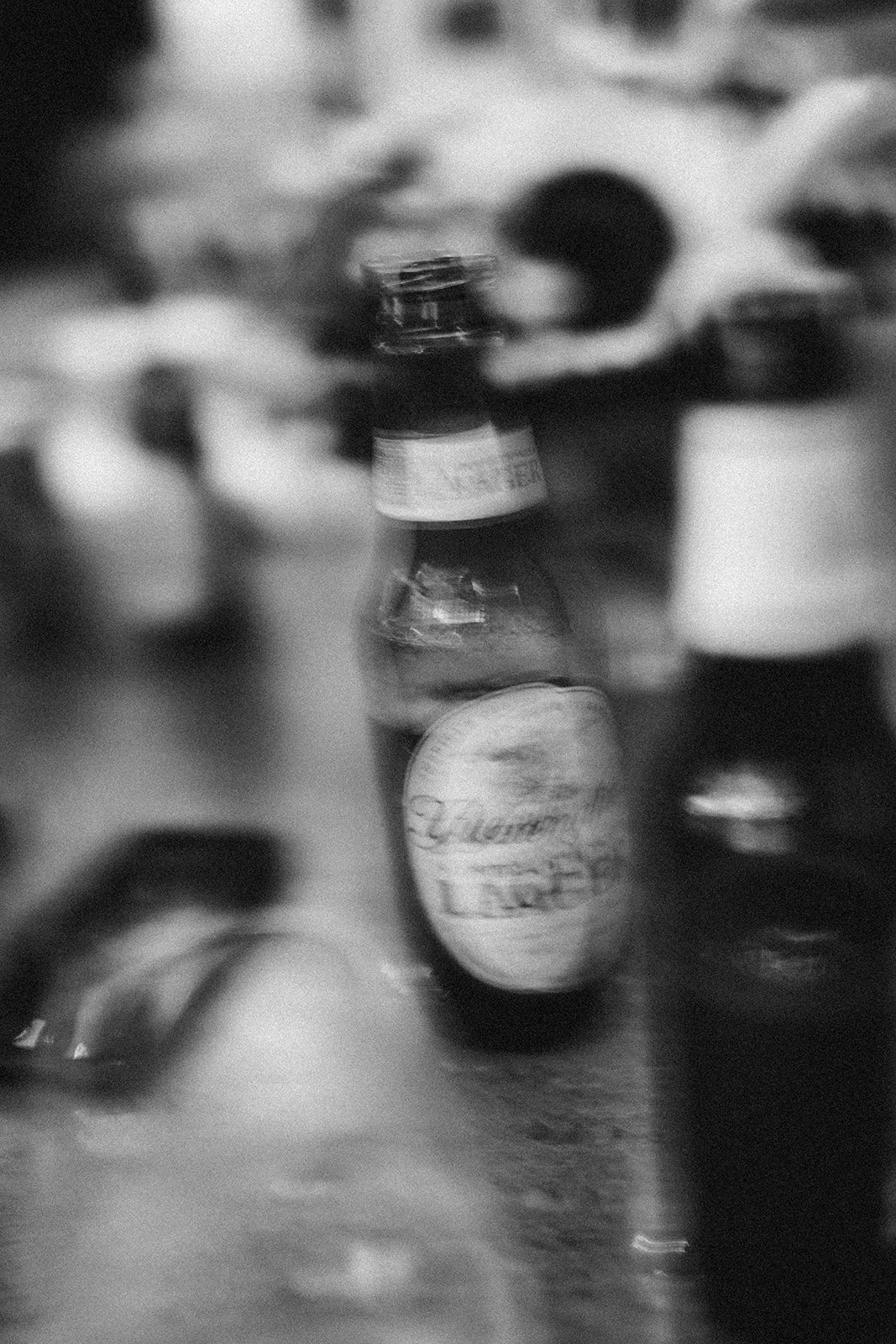 Varias botellas de diferentes formas y tamaños en un mostrador o mesa, en un entorno que parece cafetería o bar, en blanco y negro.