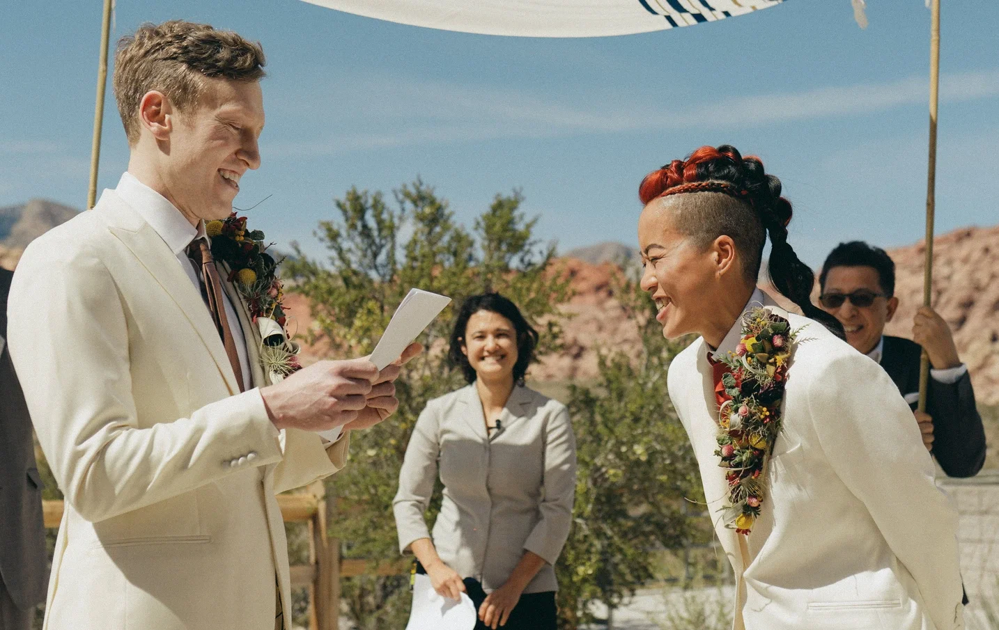Pareja en boda con trajes blancos, riendo mientras intercambian votos en un entorno al aire libre con montañas y árboles, en un día soleado.