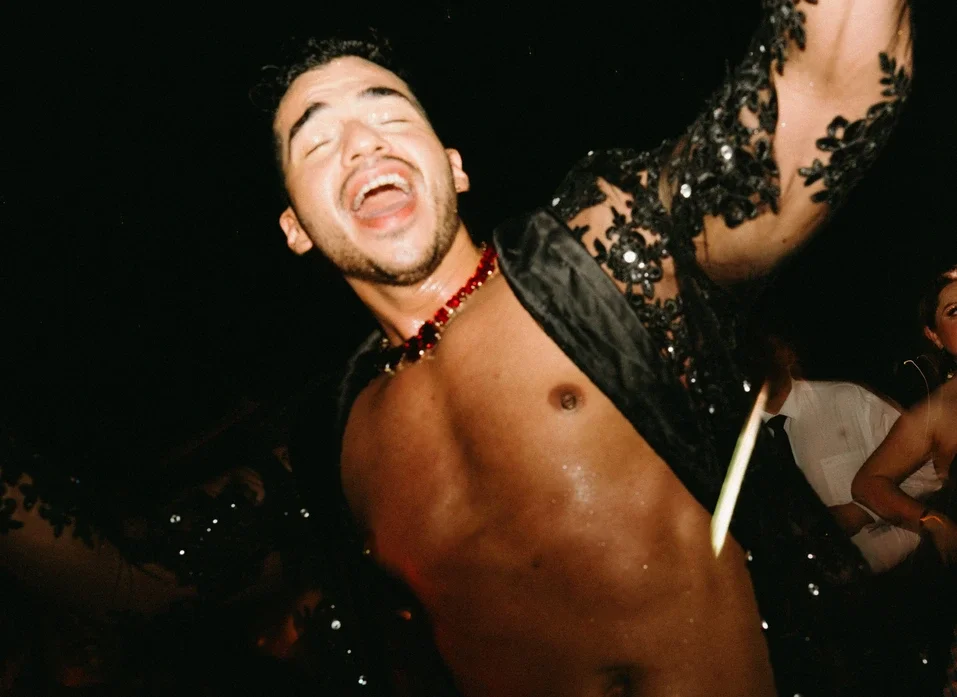 Hombre en fiesta con ropa elegante y expresión feliz, rodeado de gente.