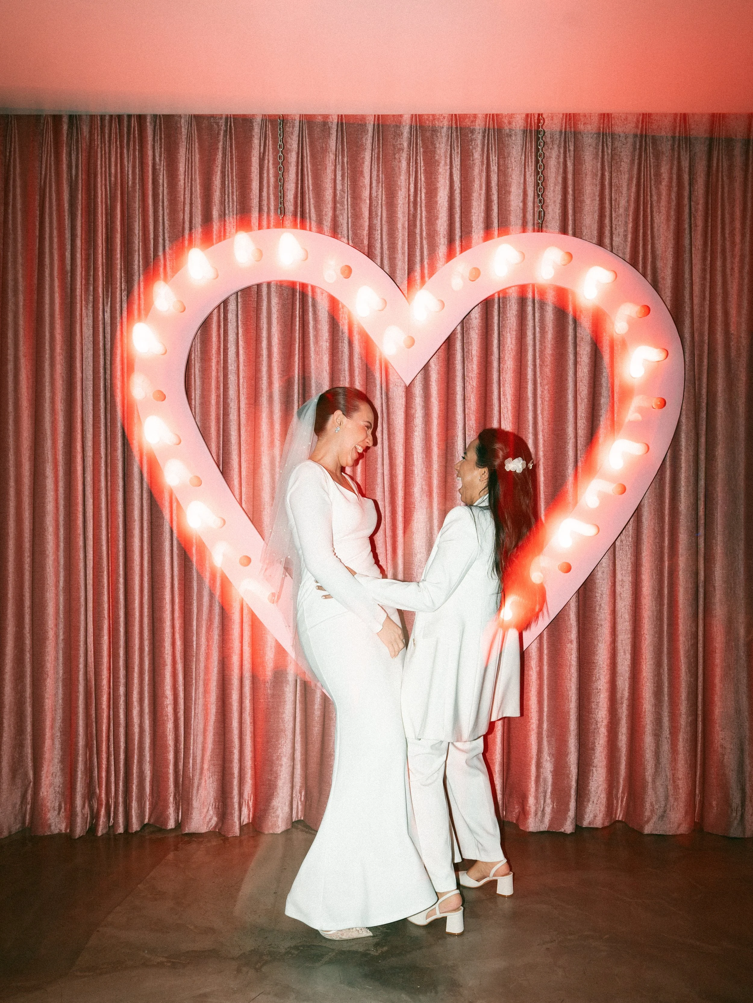 Pareja bailando en una ceremonia de boda, frente a un gran corazón con luces y cortinas rosas de fondo.