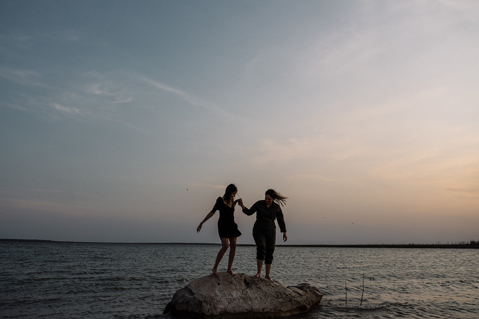 Dos mujeres caminando sobre una roca en el agua durante el atardecer, con el cielo y el mar de fondo.