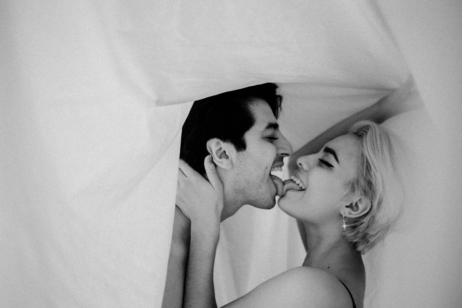Una pareja joven con expresión de alegría, tocándose la cara y con la lengua tocándose, en una pose íntima bajo una tela que forma un refugio o escondite.