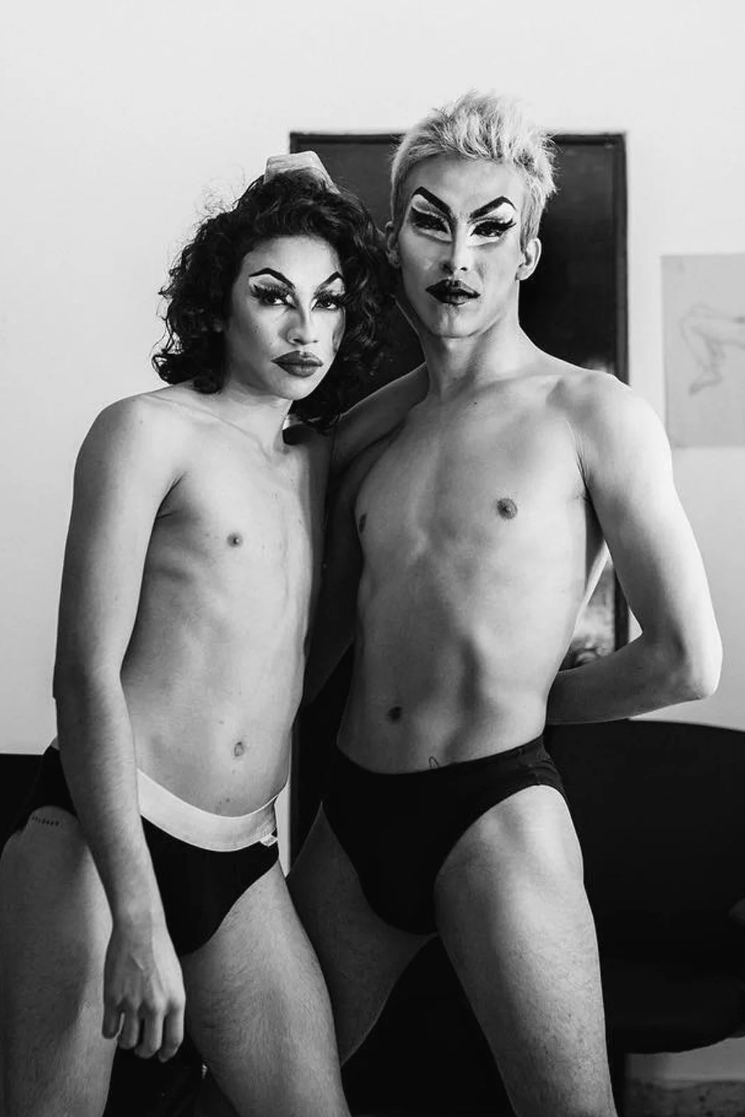 Dos personas con maquillaje llamativo posando sin ropa y con ropa interior negra en un cuarto.