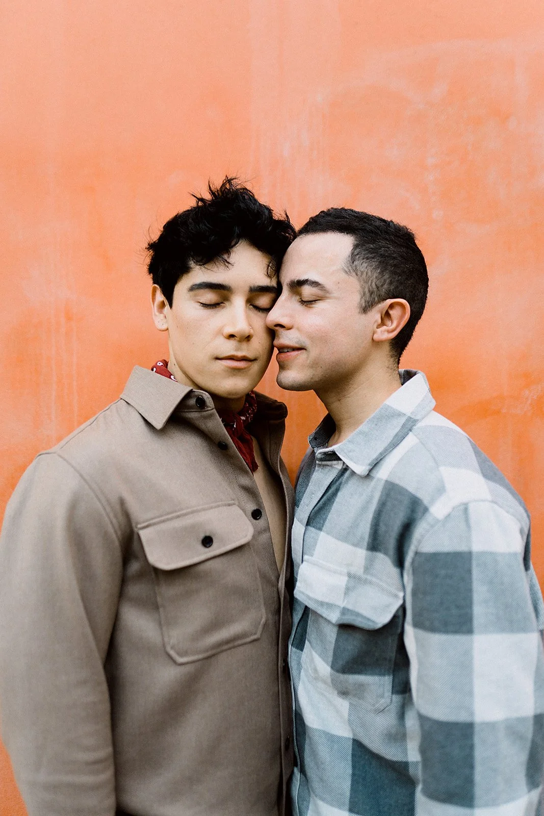 Dos hombres jóvenes con los ojos cerrados, tocándose la cara y acercándose como si compartieran un momento íntimo, frente a un fondo naranja.