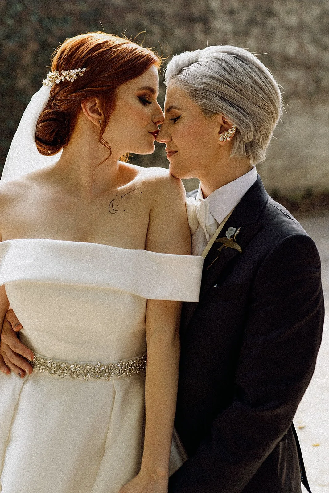 Pareja de lesbianas en boda, una con vestido de novia y la otra con traje, tocándose la frente y sonriendo. Lauros y joyas, fondo difuso.
