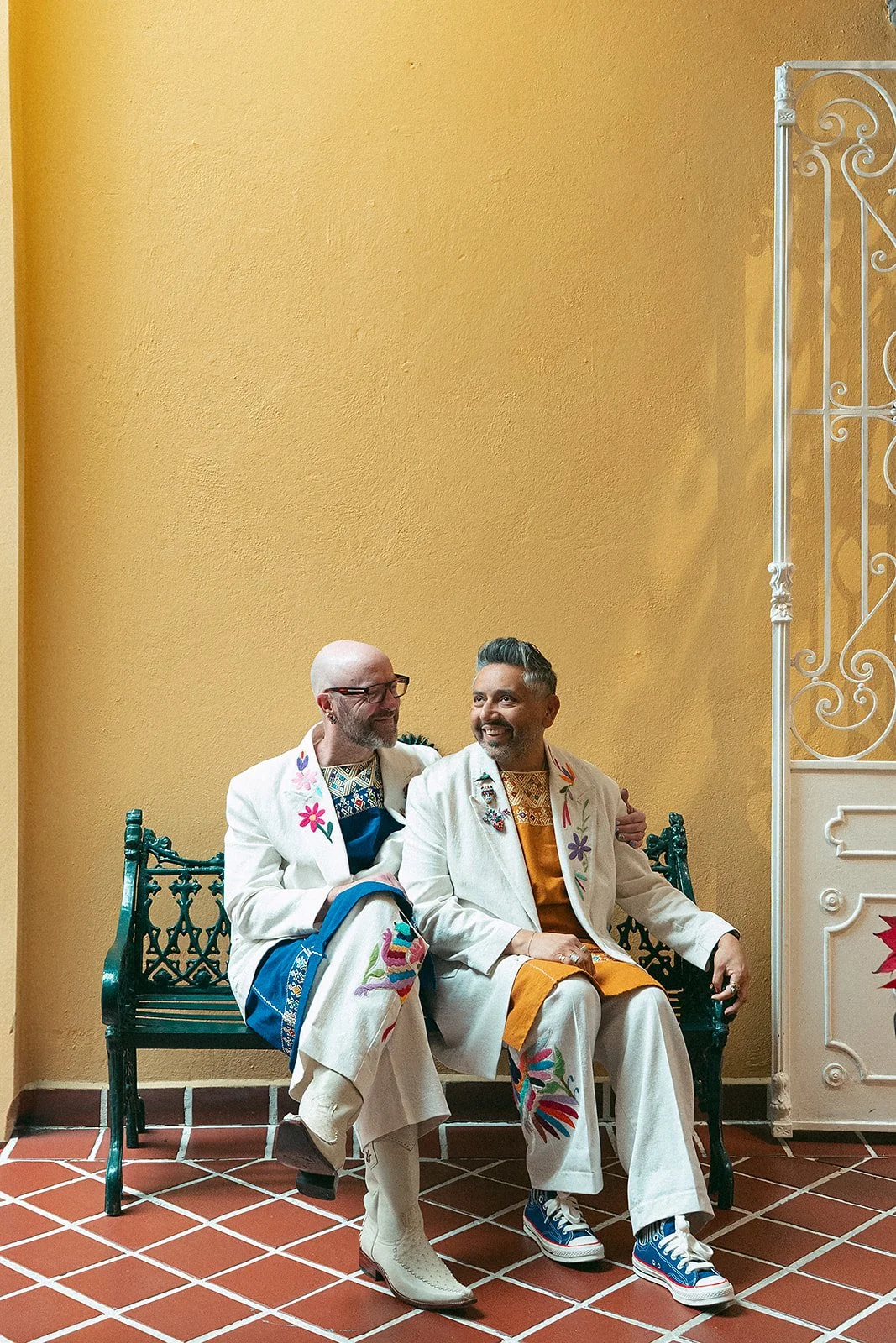 Dos hombres sentados en un banco, vestidos con ropa colorida y decorada con bordados florales, en una pared amarilla y con suelo de baldosas. Uno abraza al otro, ambos son felices.