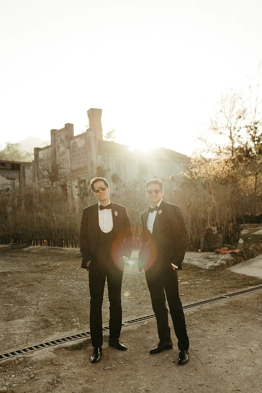 Dos hombres con trajes formales y gafas de sol en un ambiente al atardecer, de fondo un edificio antiguo y árboles.