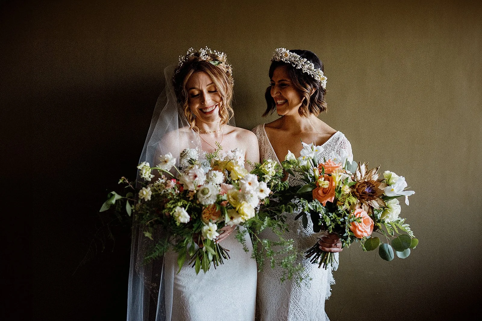 Dos mujeres en vestidos de boda sosteniendo arreglos florales, una con velo y ambas con coronas de flores, sonriendo y disfrutando del momento.