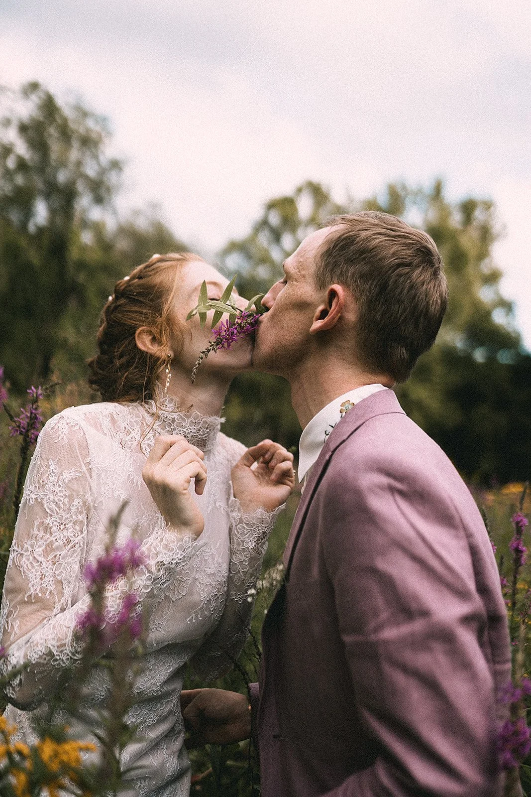 Una pareja se besa en un campo con flores, la mujer lleva un vestido de encaje y el hombre un traje rosa.