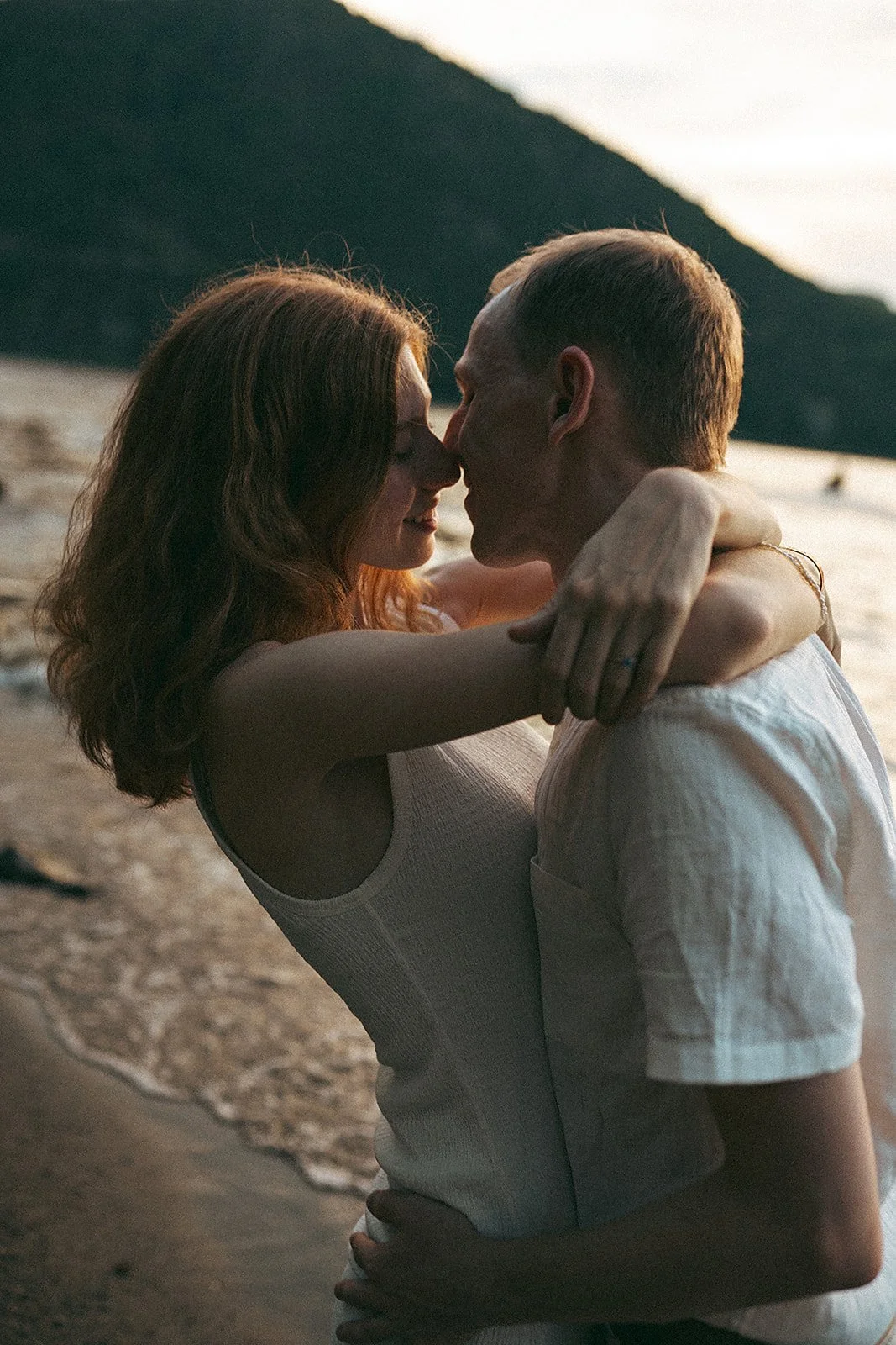 Una pareja se abraza y se acerca en la playa durante el atardecer, con un fondo de montañas y el mar.