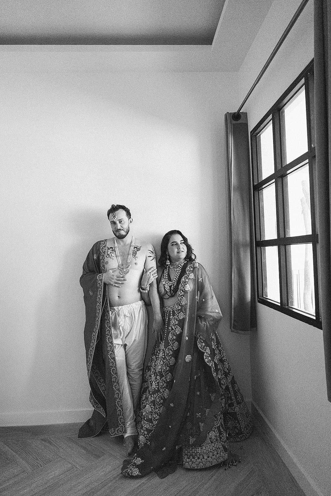Dos personas vestidas con ropa tradicional, uno con un sari y el otro con ropa de estilo antiguo, posando en una habitación junto a una ventana