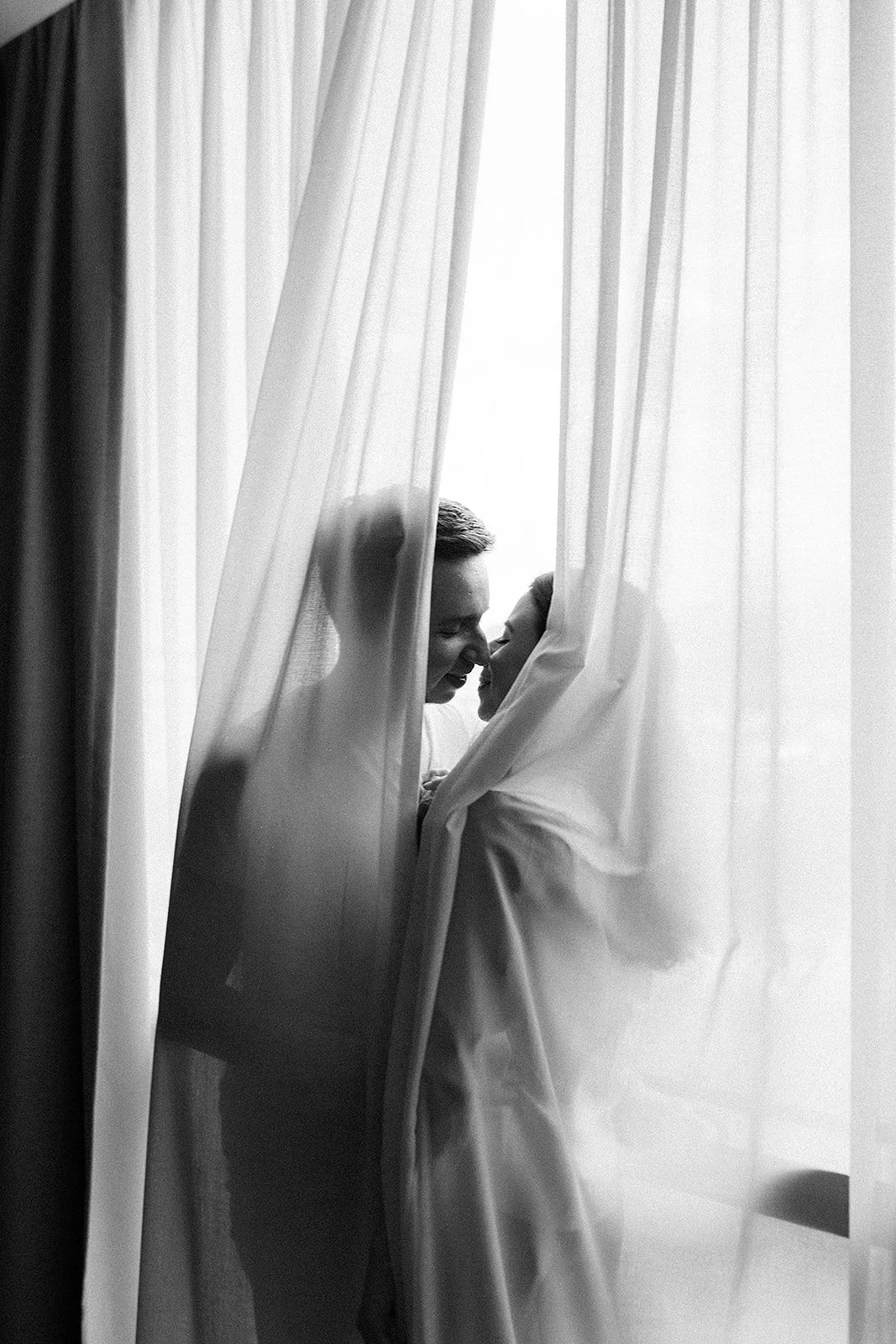 Dos personas, una adulta y una niño, con narices tocándose, entre cortinas blancas, en una escena íntima y cariñosa en blanco y negro.