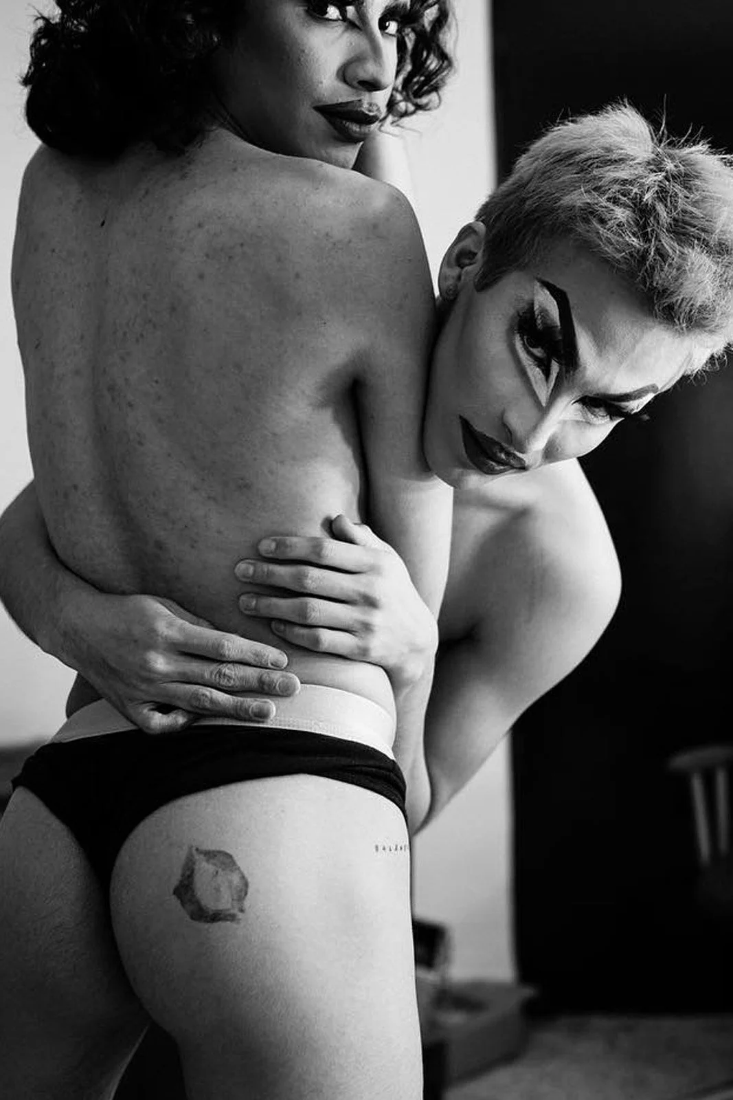 Dos mujeres de cabello rizado y corto posando juntas en una fotografía en blanco y negro. Una de ellas tiene tatuaje en la parte superior del muslo.