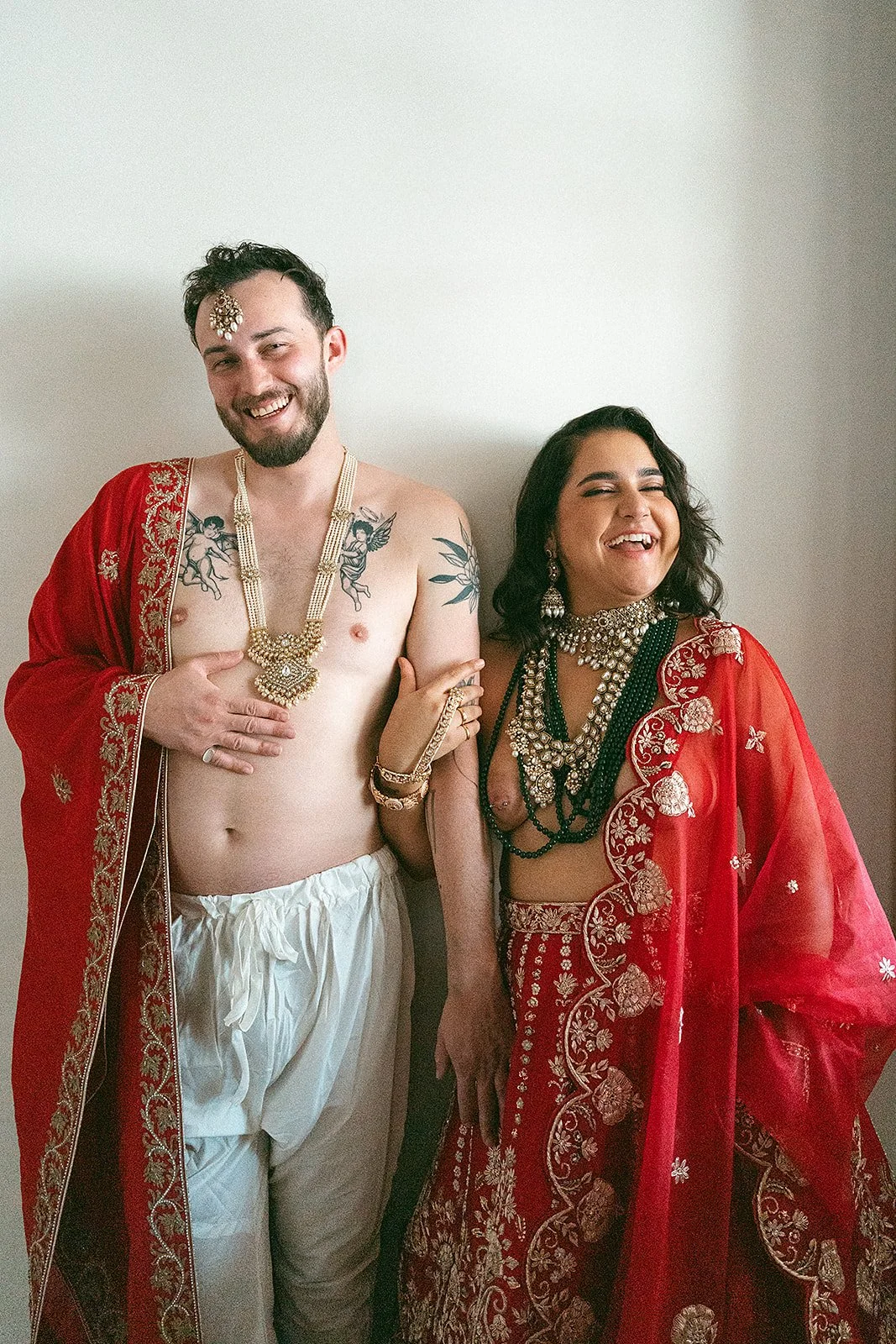 Dos personas sonriendo y posando con trajes tradicionales indios, la persona a la izquierda es un hombre sin camisa con accesorios dorados, y la persona a la derecha es una mujer con un sari rojo y joyería.