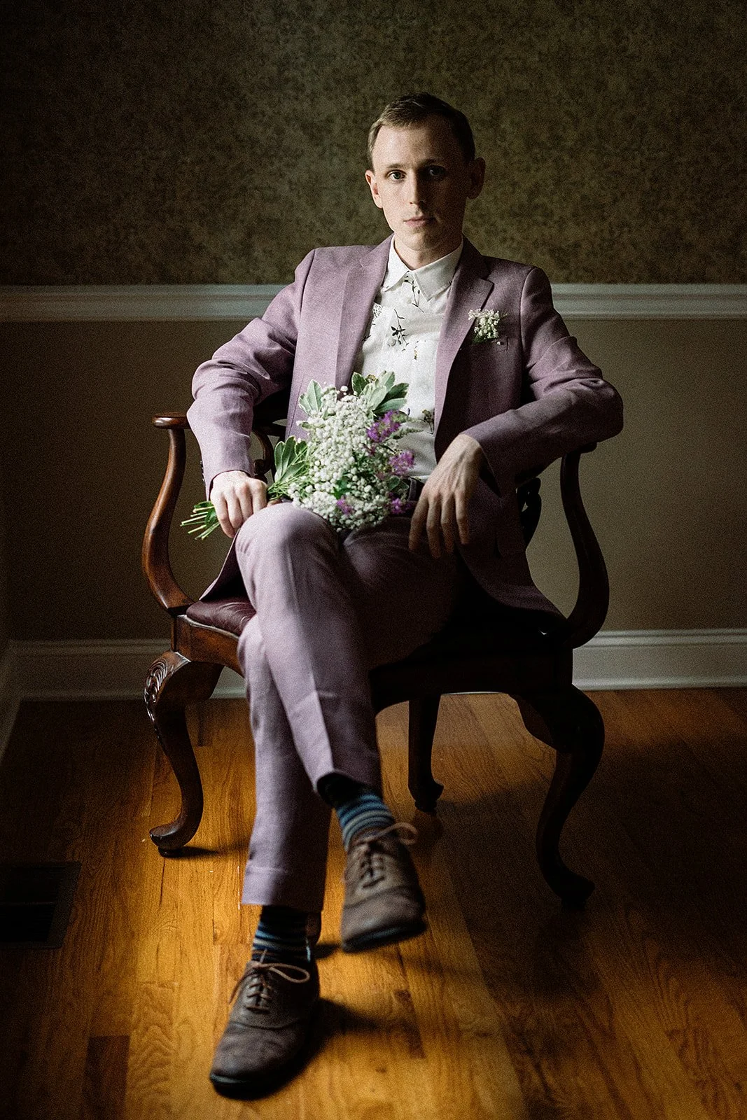 Hombre joven sentado en una silla de madera, vestido con saco y pantalón rosas, sosteniendo un ramo de flores en su regazo, en una habitación con paredes decoradas y suelo de madera.