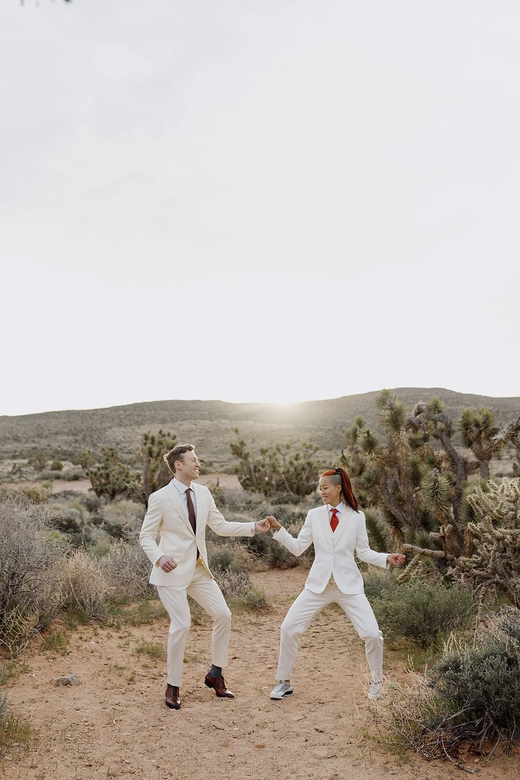 Una pareja bailando en un desierto con cactus, ambos vestidos con trajes blancos y faldas, con el sol saliendo o poniéndose en el horizonte.