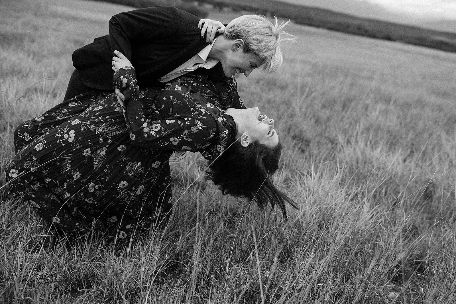 Pareja en un campo abierto, con la mujer inclinandose hacia atrás y el hombre sosteniéndola, ambos sonriendo y disfrutando del momento.