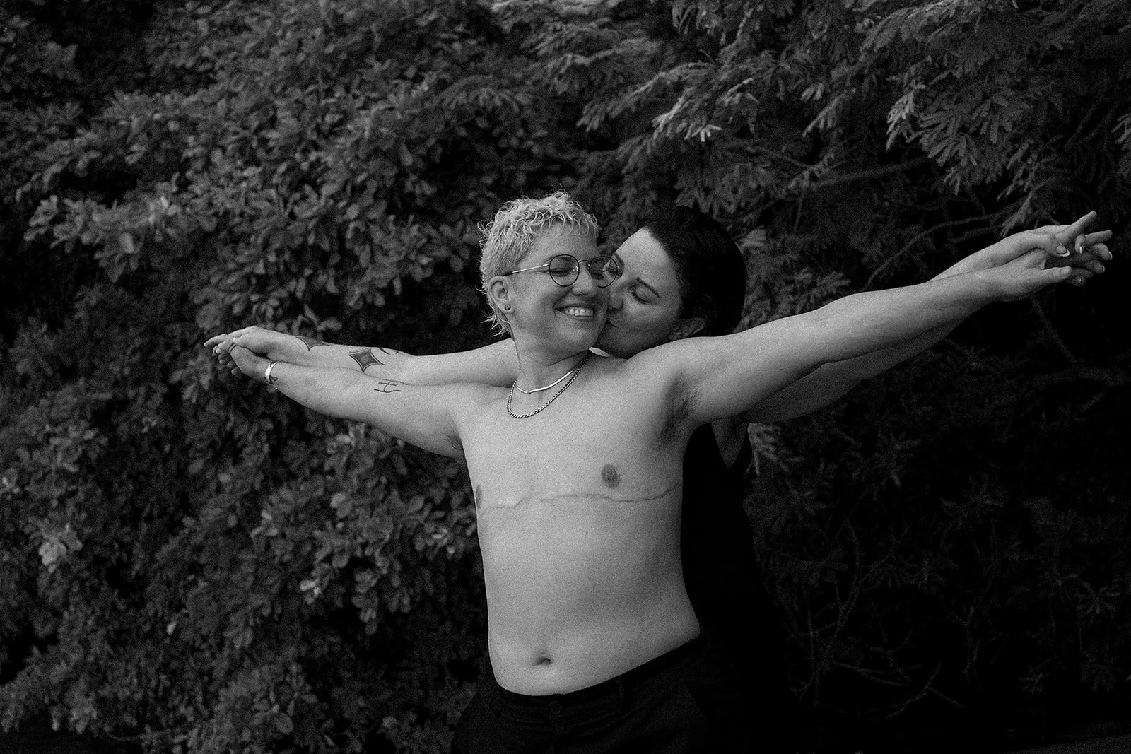Dos personas desnudas disfrutando de un momento de alegría, abrazadas en la naturaleza, una de ellas con cabello rubio, gafas y tatuajes, la otra con cabello oscuro y en saludo cariñoso, rodeadas de hojas.