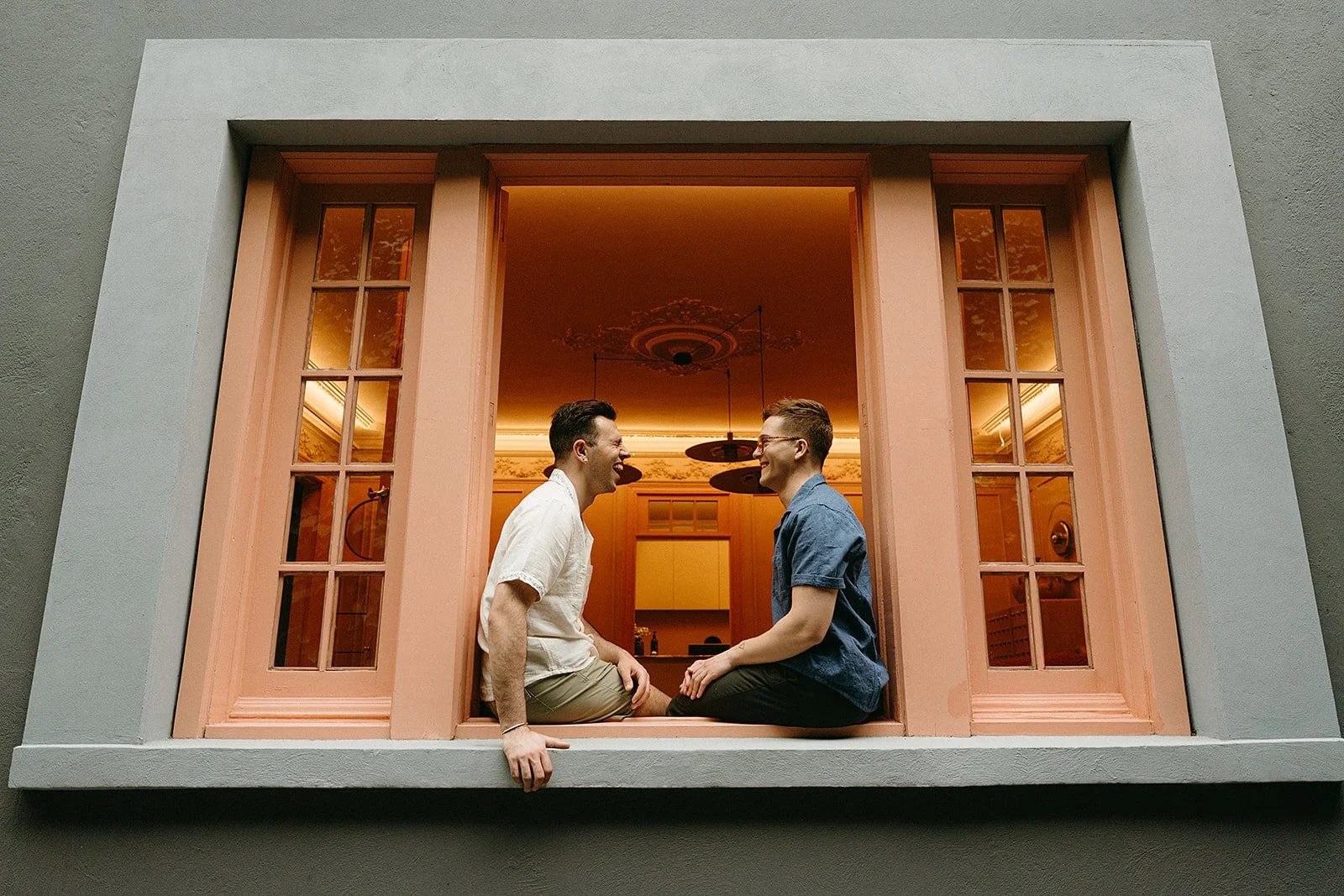 Dos hombres jóvenes riendo y sosteniéndose de las manos, sentados en una ventana abierta en un apartamento elegante, con decoraciones clásicas y techos altos.