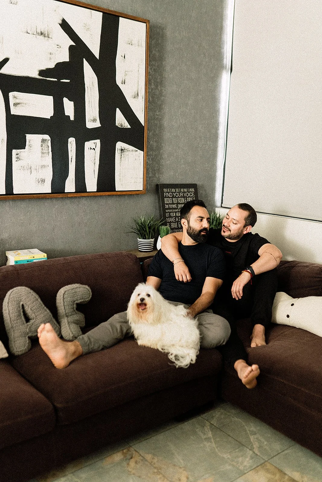 Dos hombres sentados en un sofá marrón, uno con un perro blanco y esponjoso en sus piernas. Uno abraza al otro y ambos parecen conversar. En el fondo, cuadros y plantas decorativas en una sala moderna.