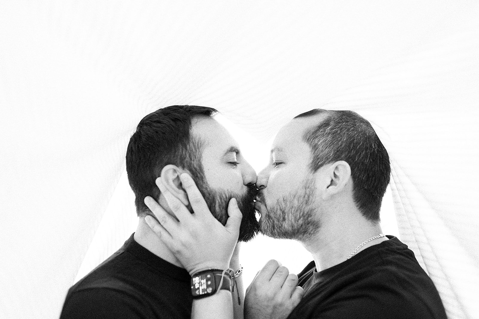 Dos hombres con barba se besan en la frente, tocándose las caras con las manos, en un fondo blanco