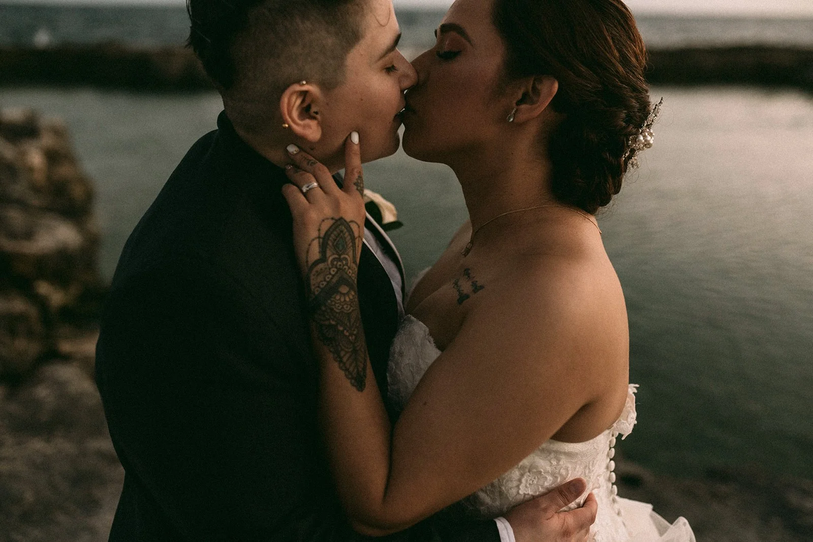 Una pareja se besa en la playa al atardecer, ambos vestidos de manera formal, ella con vestido de novia y él con traje. La mujer tiene tatuajes y peinado elaborado con accesorios.