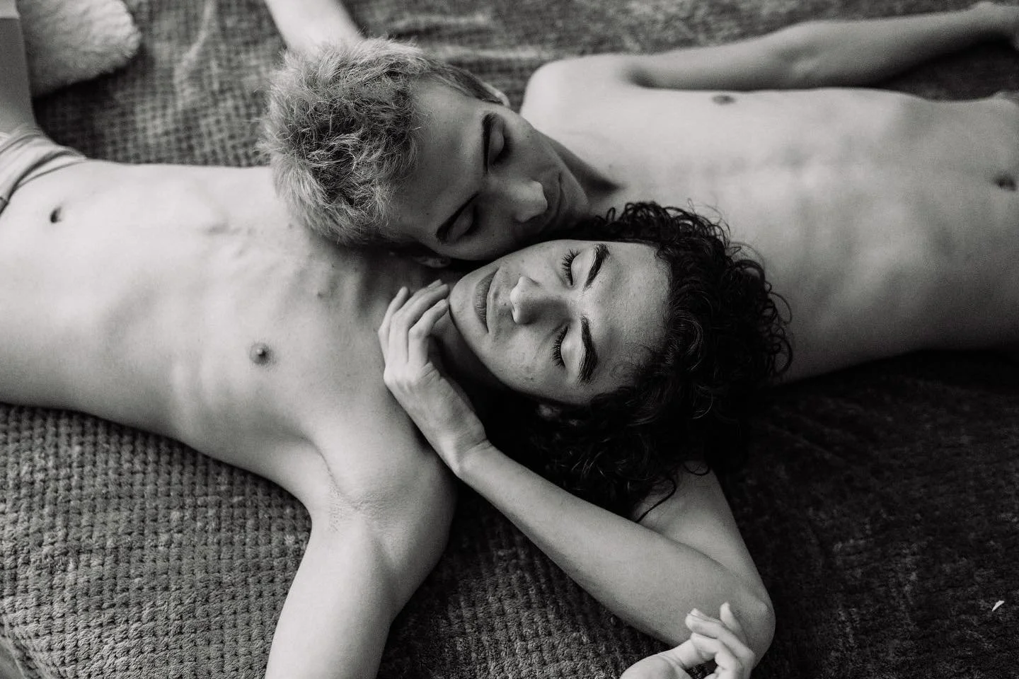 Dos personas desnudas, una con cabello rubio y otra con cabello rizado negro, descansando juntas con los ojos cerrados sobre una superficie texturizada.