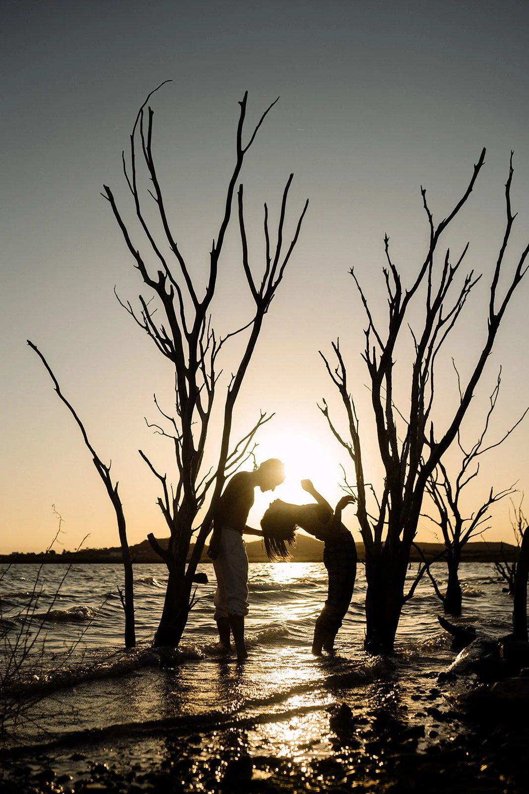 Dos personas en un río durante el atardecer, entre árboles secos, una tocando la cabeza de la otra.