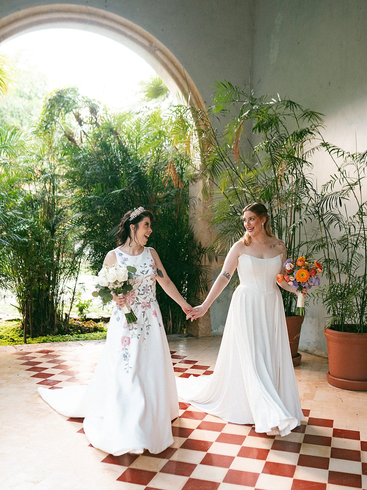 Dos novias lesbianas en boda, una con vestido de novia sin mangas y otra con vestido de tirantes, sosteniéndose de la mano y sonriendo, rodeadas de plantas verdes y luz natural.