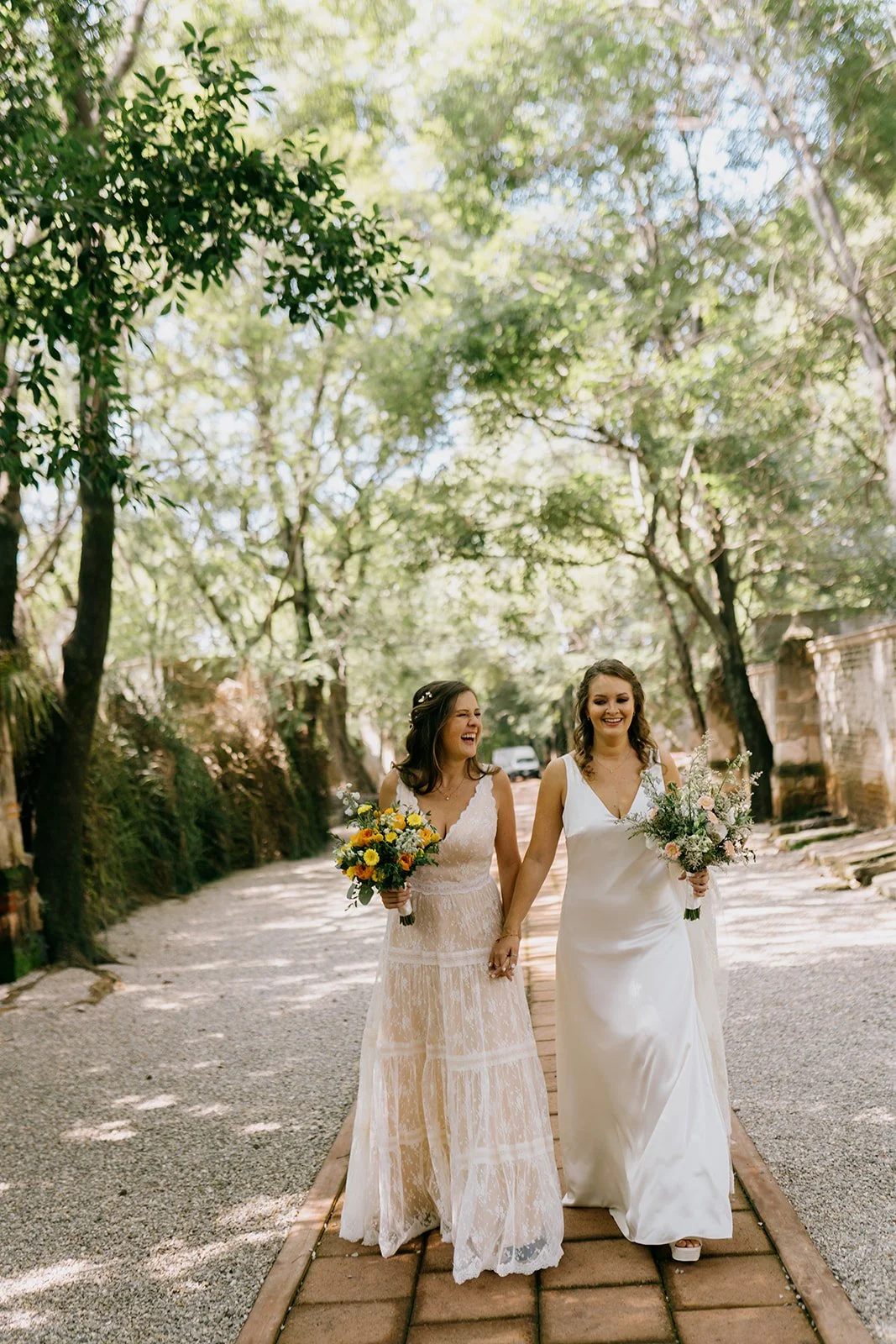 Dos mujeres vestidas con vestidos de novia, caminando juntas por un sendero en un parque, sonriendo y sosteniendo ramos de flores.
