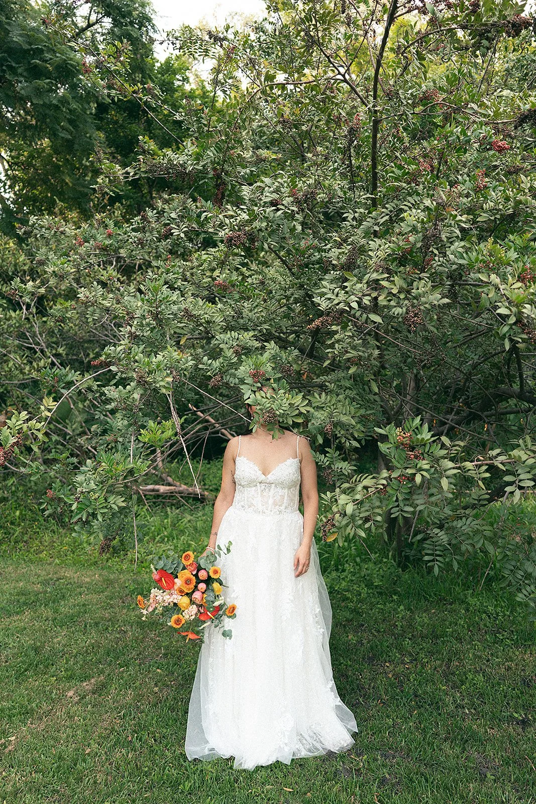 Una mujer con vestido de novia sostiene un ramo de flores y se encuentra en un jardín con árboles frondosos y césped.