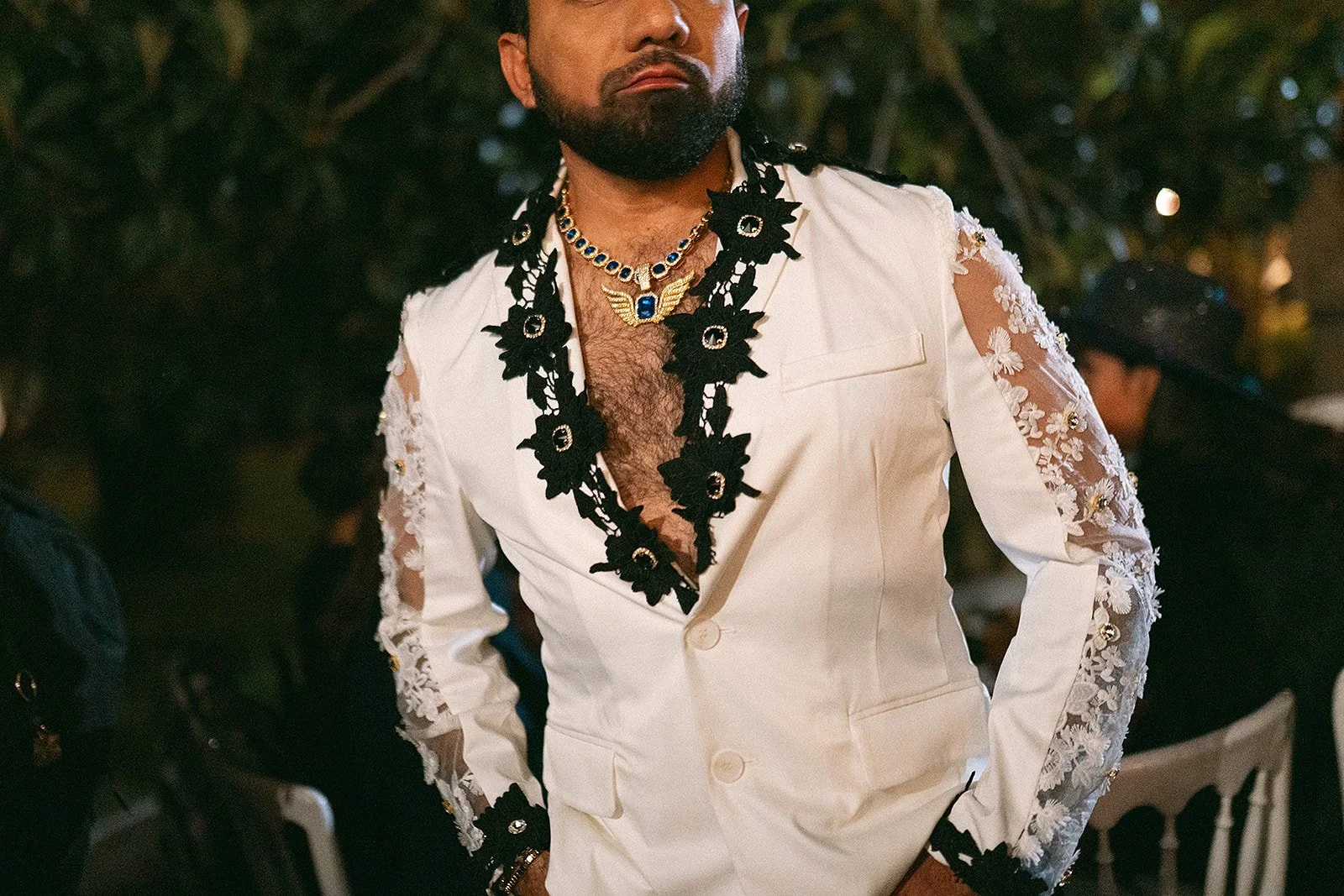 Hombre con barba y cabello largo, vestido con chaqueta blanca con detalles de encaje y accesorios negros con flores, en un evento social por la noche.