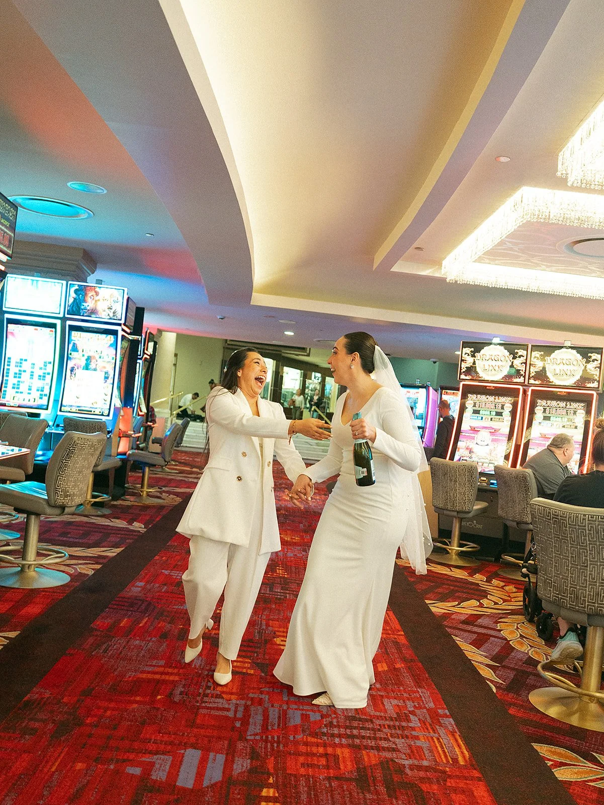 Dos mujeres vestidas de blanco en una sala de tragamonedas, una de ellas con un ramo de flores y la otra con una botella, parecen felices y celebrando.