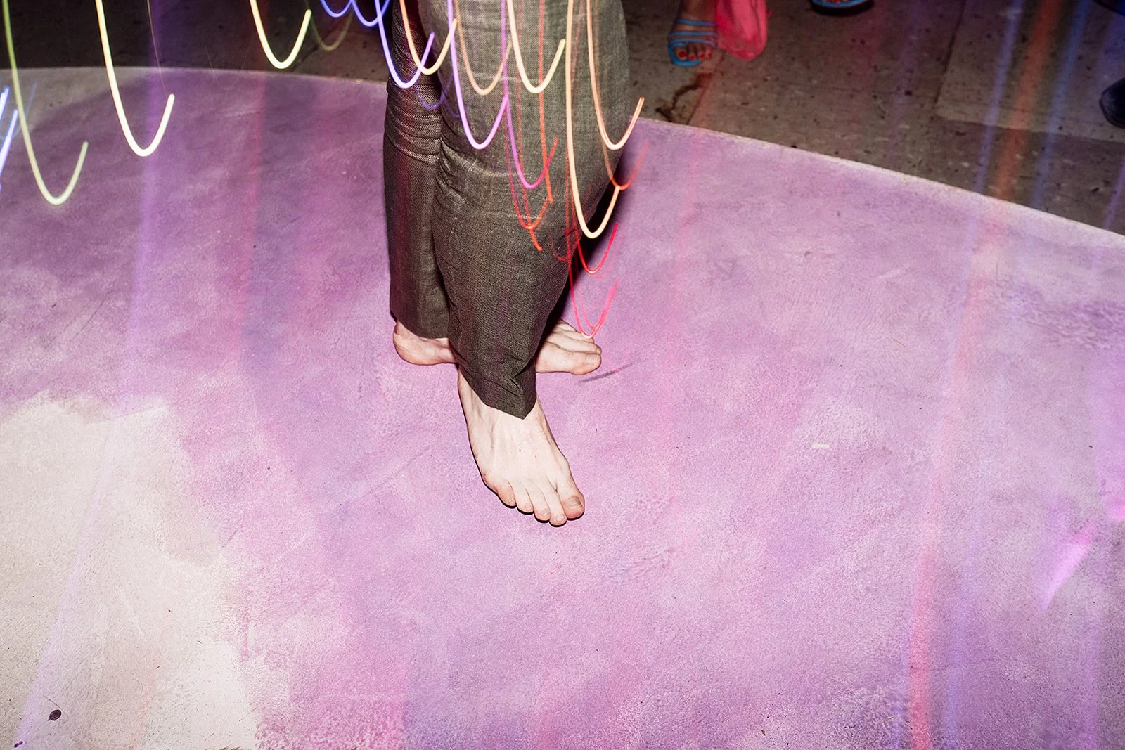 Persona con pantalones de rayas, descalza, de pie sobre una superficie rosa, con algunas luces de neón y una superficie de piso de cemento en el fondo.