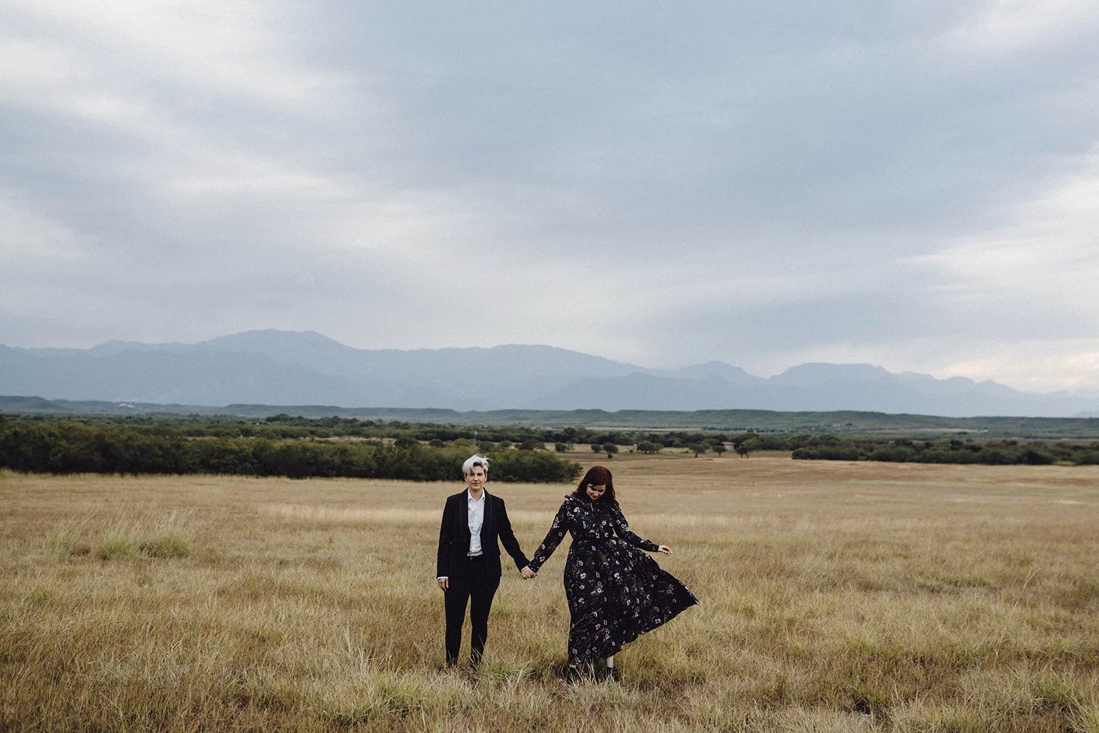 Dos personas caminando juntas por un campo con montañas al fondo, una de ellas con vestido largo y la otra con saco y pantalones, en un día nublado.