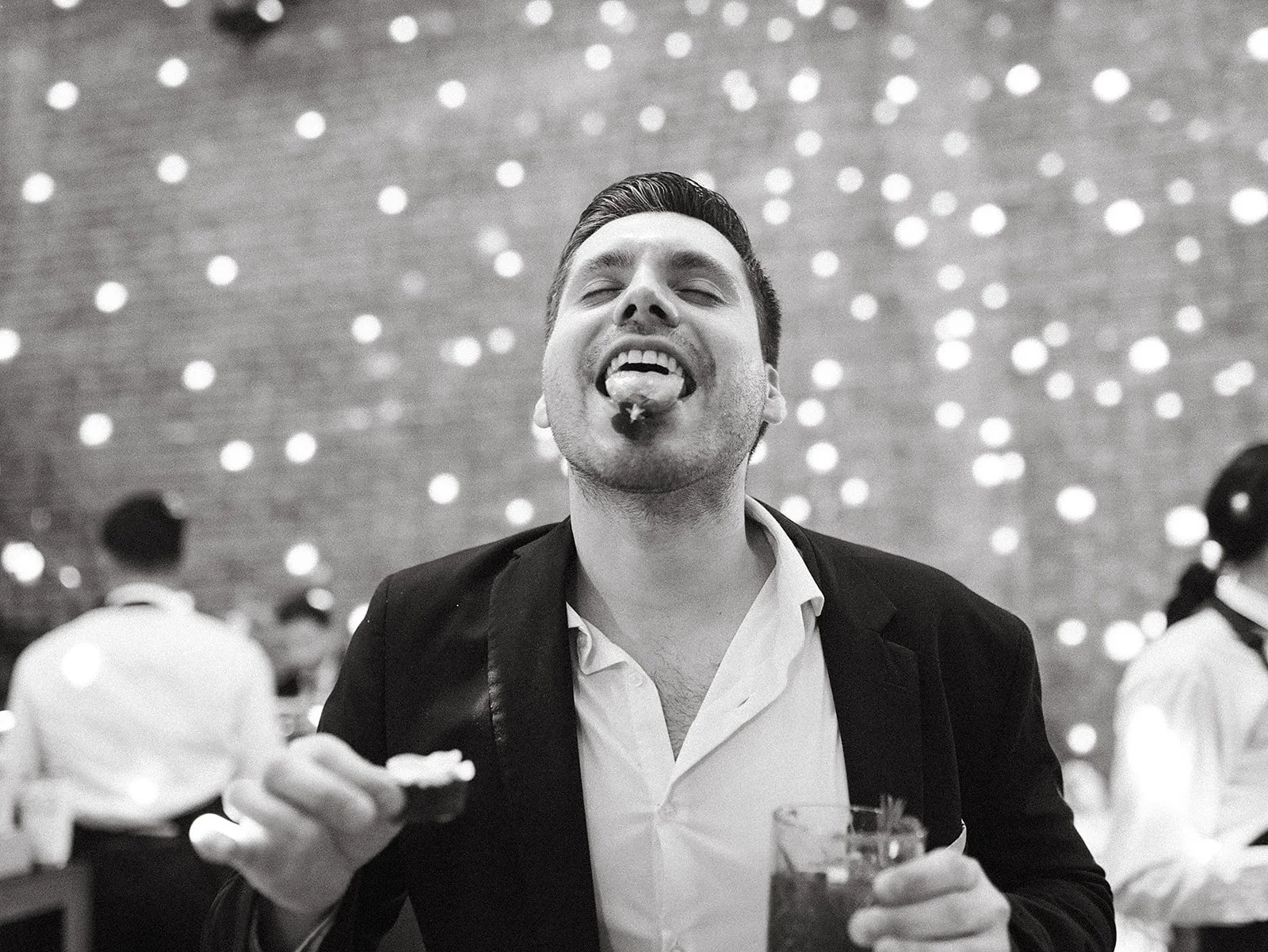 Hombre en un evento social, sonriendo, con una bebida en cada mano, fondo con luces decorativas, en blanco y negro.