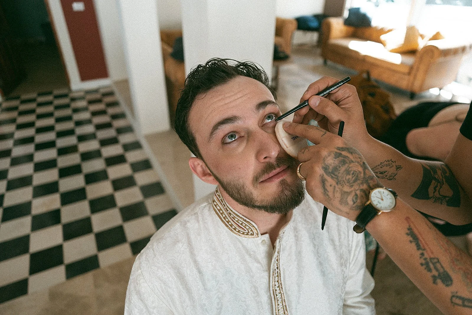Hombre con barba, de cabello oscuro, sentado en una sala, recibiendo maquillaje con un pequeño pincel y siempe con los ojos mirando hacia arriba.