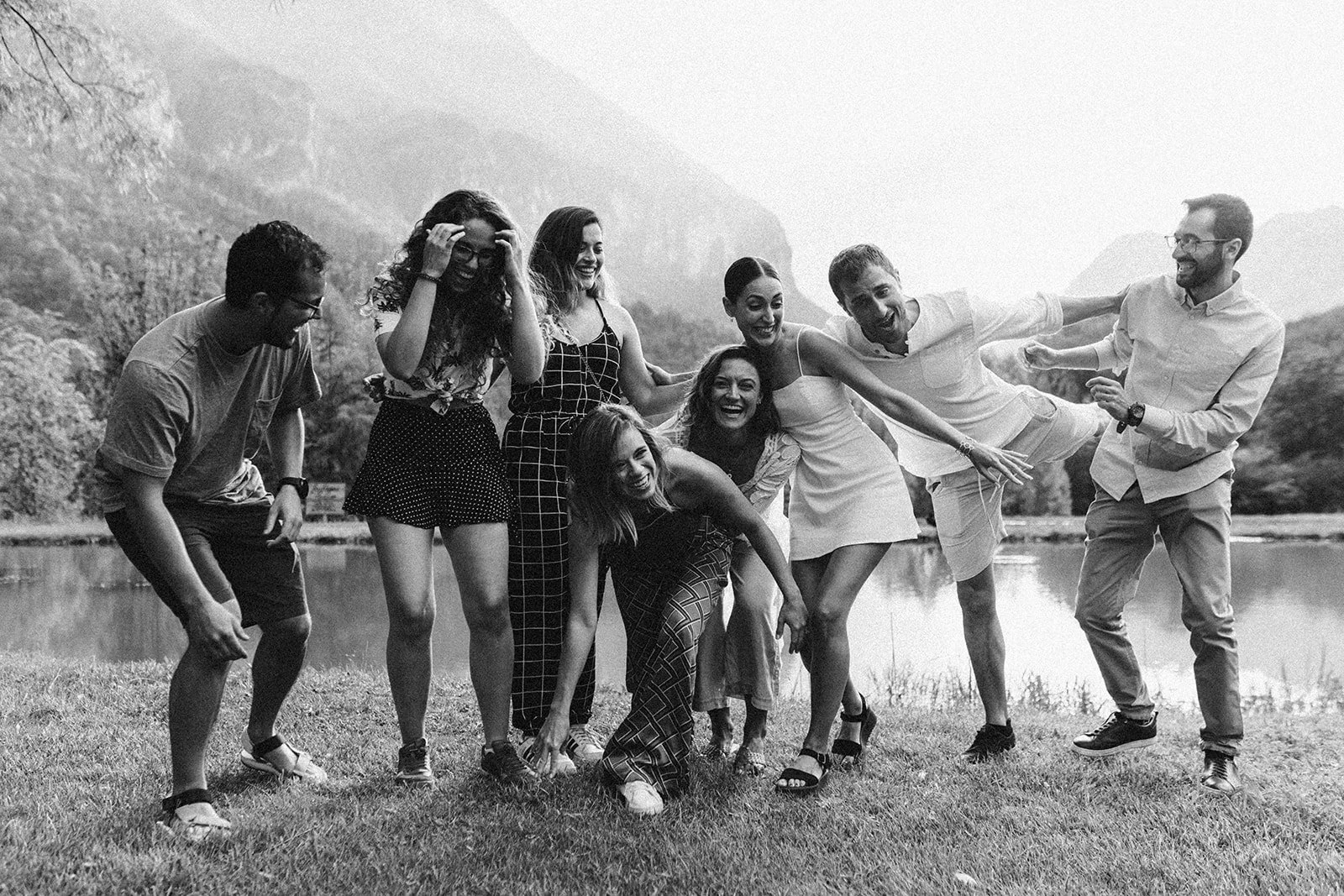 Grupo de personas riendo y jugando al aire libre cerca de un lago con montañas al fondo, en blanco y negro.