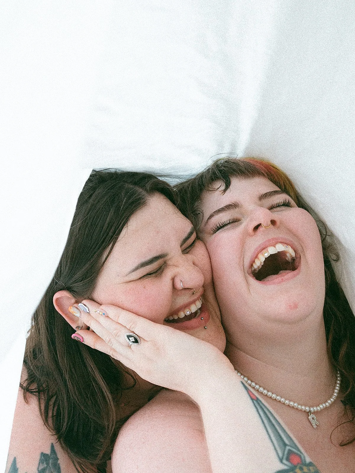 Dos jóvenes sonriendo y riendo juntas, con una en el centro y la otra tocándose la cara, en un fondo blanco.