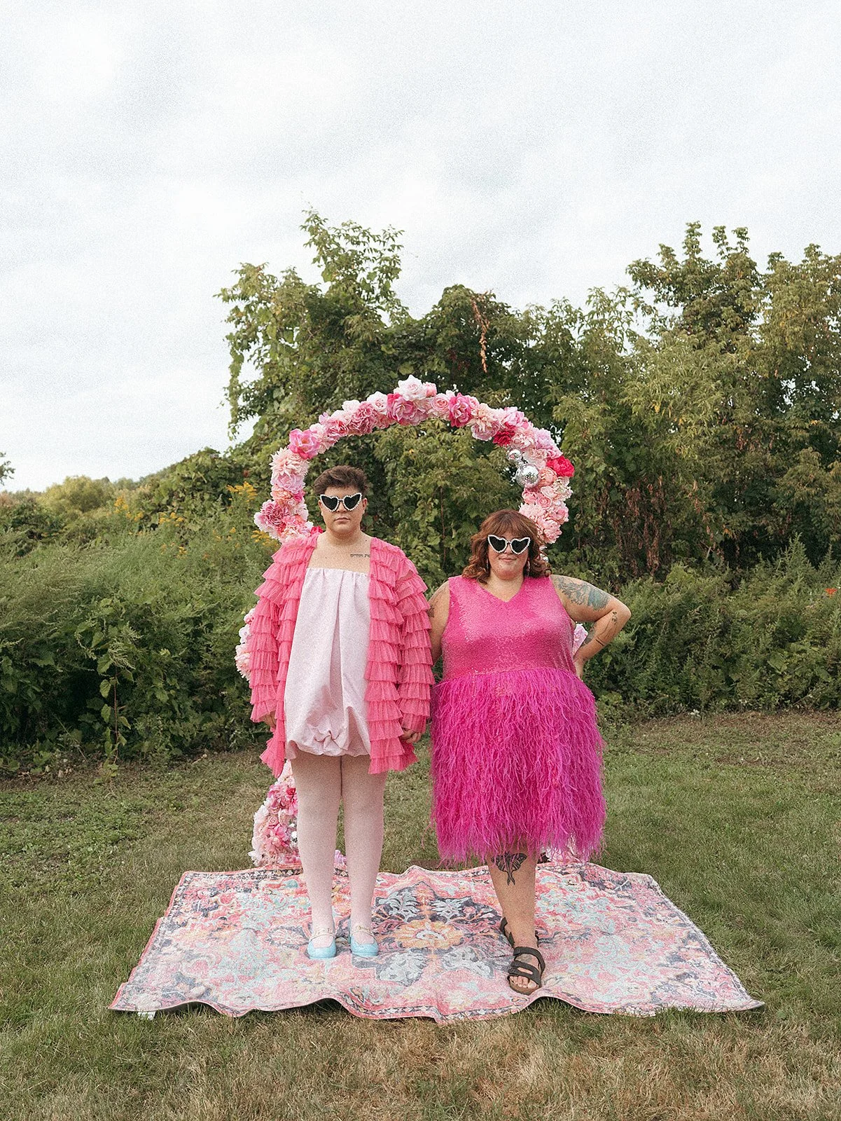 Dos personas con gafas de sol de corazón, vestidas con ropa rosa, posando frente a un arco de flores rosadas en un campo verde.