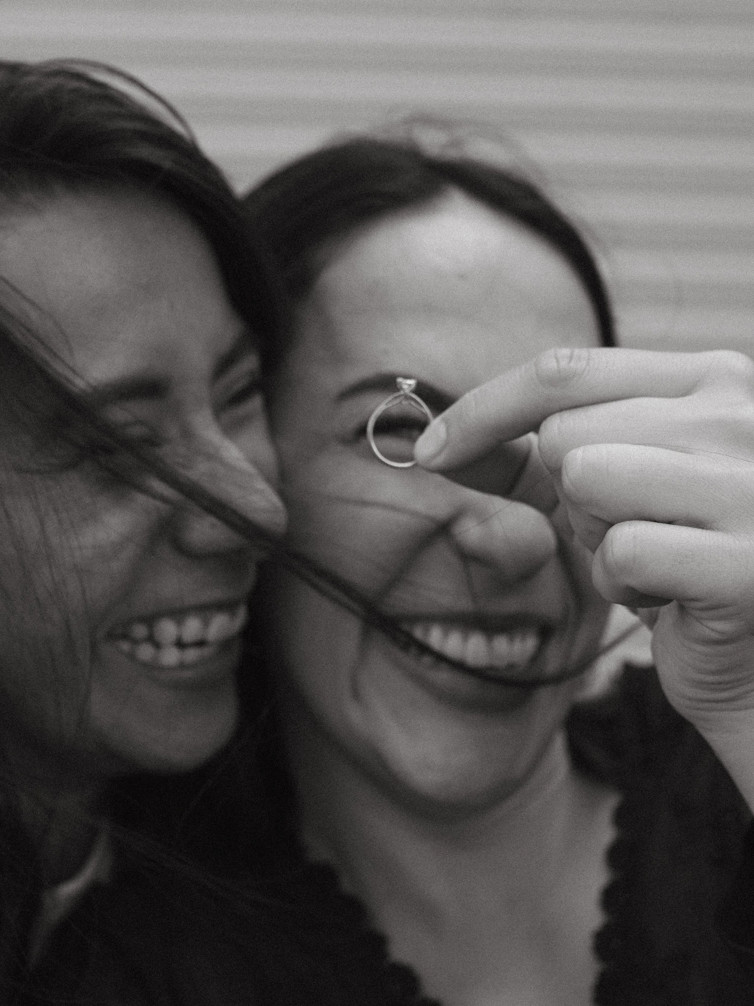 Dos personas sonriendo y tocándose frente a una cámara, una lleva un pendiente y la otra tiene un anillo en el dedo índice, en un ambiente de camaradería.