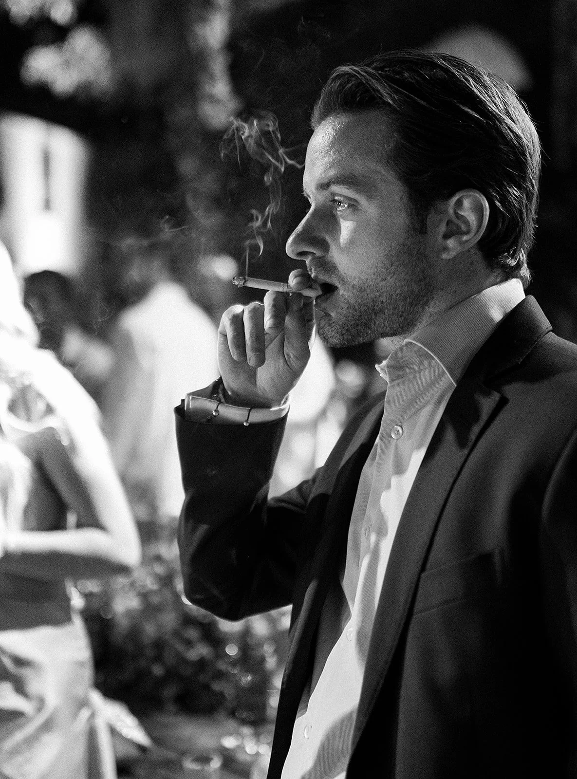 Joven hombre con cabello peinado, vestido con traje formal y camisa, fuma un cigarrillo en un ambiente social nocturno, con otras personas en el fondo.