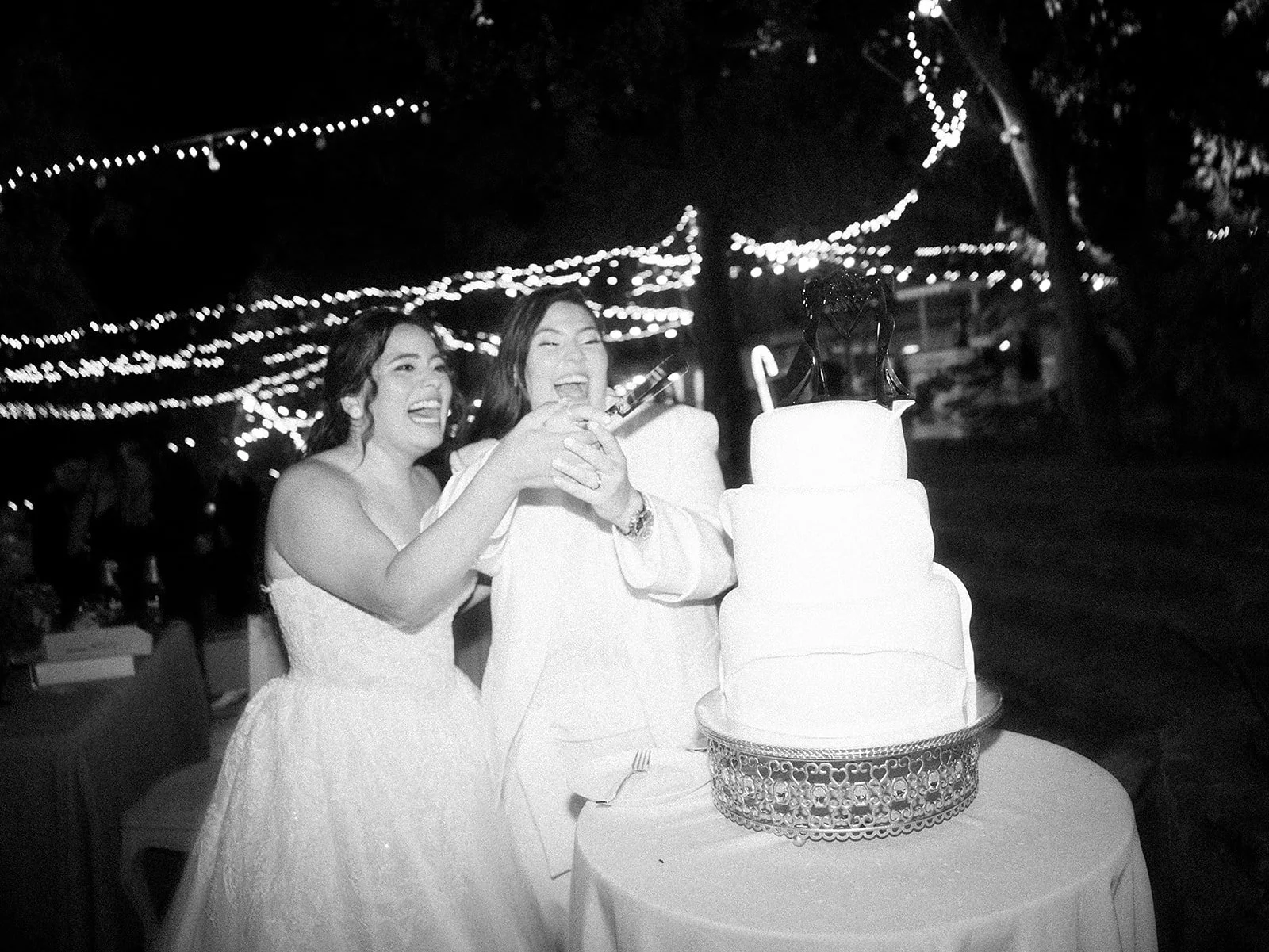 Pareja de novios en su boda cortando un pastel de bodas, rodeados de luces decorativas en un ambiente nocturno.