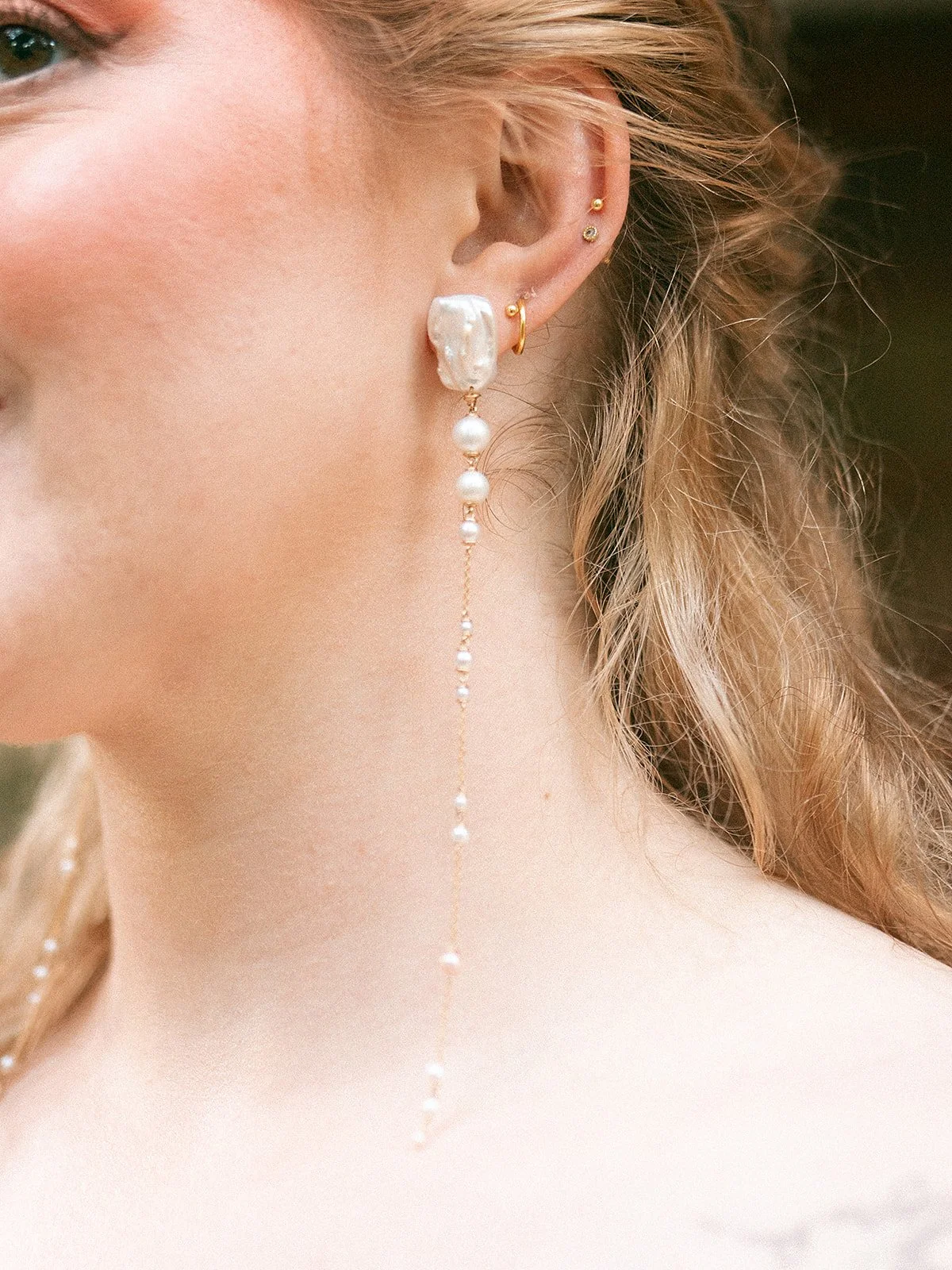Cerca de la oreja de una mujer, se pueden ver varios piercings y unos pendientes largos de perlas y oro.