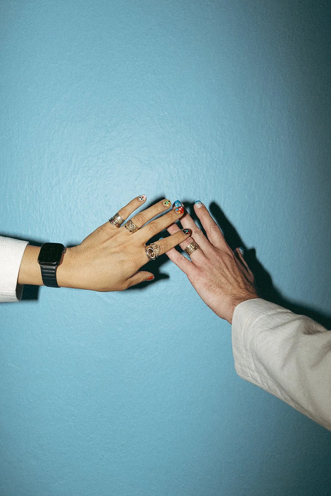 Dos manos tocándose, una con uñas decoradas de colores vibrantes y varias anillos, con reloj inteligente en la muñeca, otra con uñas pintadas y varias sortijas, contra una pared azul.