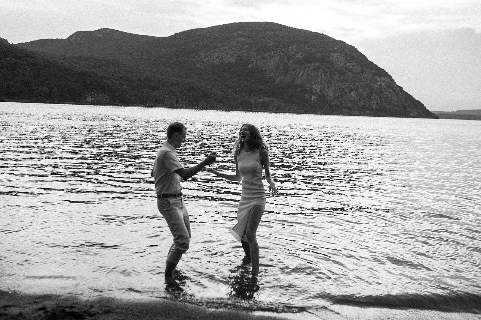 Dos personas, un hombre y una mujer, bailando y sonriendo en la orilla de un lago con montañas en el fondo.
