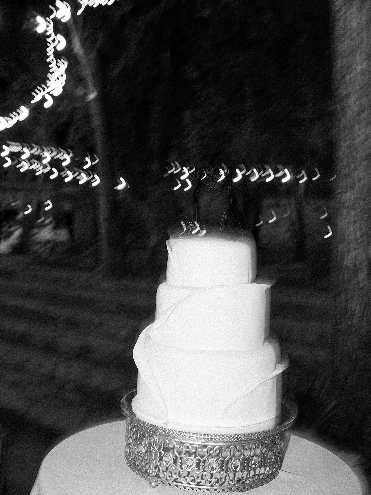 Tarta de tres pisos en blanco, decorada con papel arabesco en la base de plata, sobre una mesa plateada, con fondo oscuro y luces difusas.