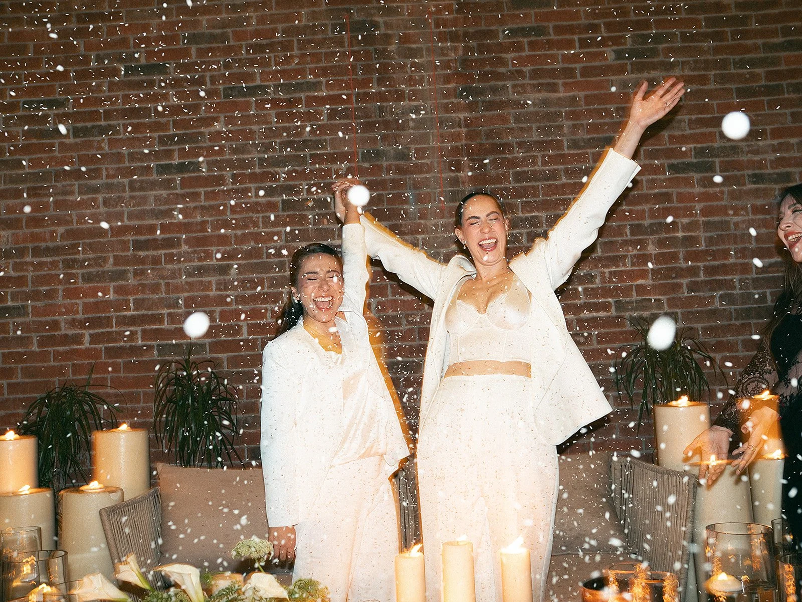 Tres mujeres riendo y celebrando en una fiesta con velas y decoraciones, lanzando confeti o nieve artificial, con un fondo de pared de ladrillos.