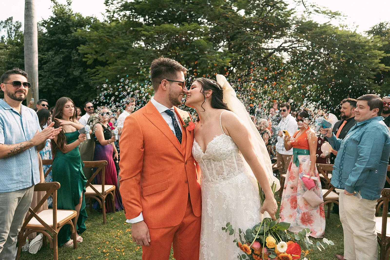 Una pareja de recién casados, un hombre con traje naranja y una mujer con vestido blanco, se besan en una boda al aire libre, rodeados de invitados que lanzan burbujas y celebran.
