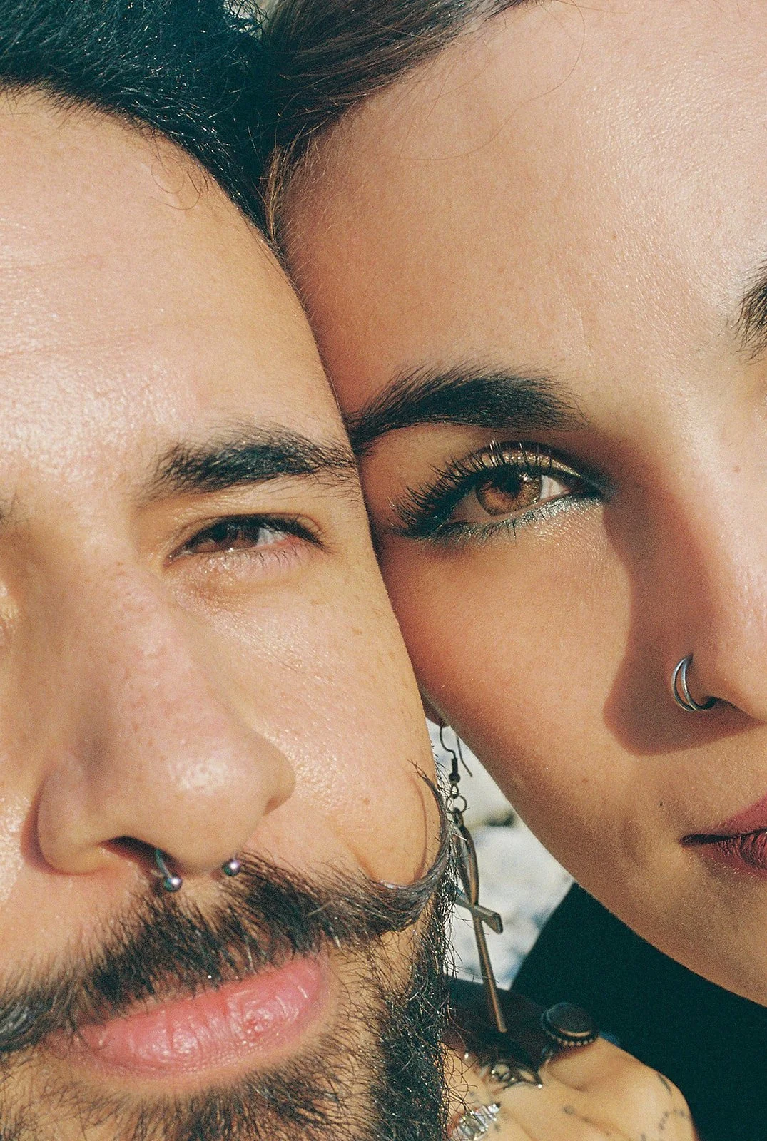 En la imagen hay una pareja con cerca de la cara, parecen estar muy juntos. La mujer tiene cejas oscuras y cabello oscuro, usa maquillaje y un aro en la nariz. El hombre tiene barba y bigote, usa piercings y accesorios en la oreja.